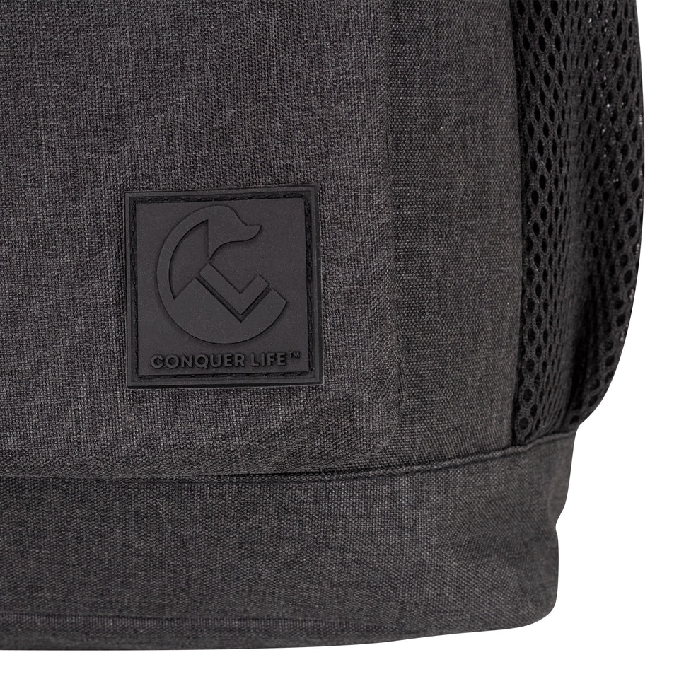 Closeup front view of black poly-fabric backpack, lower right corner, black plastic Conquer Life logo patch, 1" square